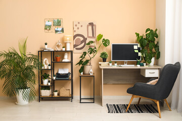 Stylish workplace with computer, shelving unit and different houseplants near beige wall at home