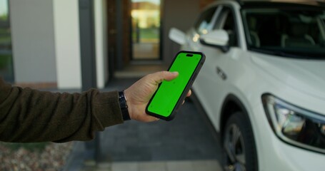 A man uses a mobile phone with green screen standing near an electric car. Close-up of his hands.