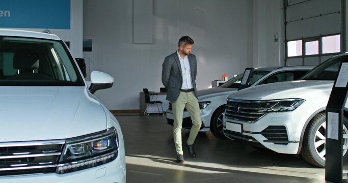 A stylish man chooses a car at a car dealership. He walks between the cars and examines them with a serious look