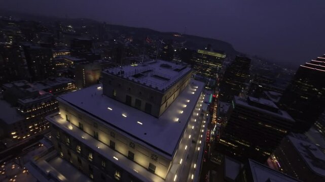 FPV drone flies above snowy roof of Sun Life Building, breathtaking night views of Square Dorchester and Montreal Downtown in winter.