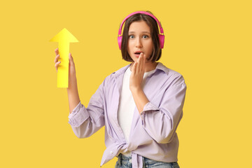 Shocked young woman in headphones with paper arrow pointing at something on yellow background