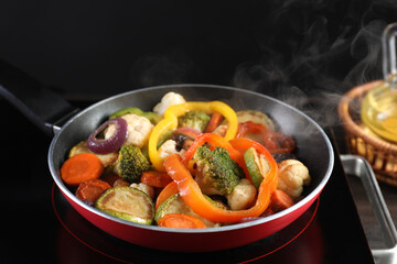 Frying pan with vegetables and stove on table against black background, closeup