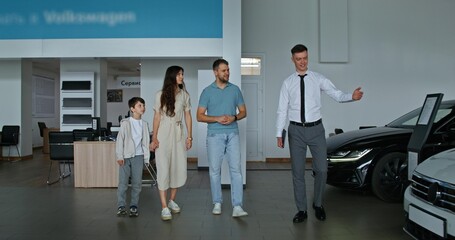Obraz premium The car dealership manager shows cars to customers by taking them through the showroom. A family with a child chooses a car