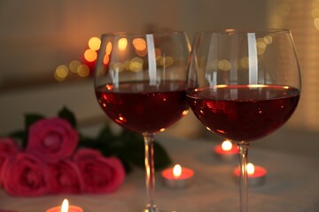 Romantic place setting with red wine, burning candles and roses on table against blurred lights,...