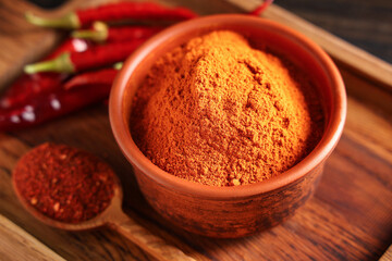 Bowl and spoon of red chili powder with peppers on wooden background, closeup