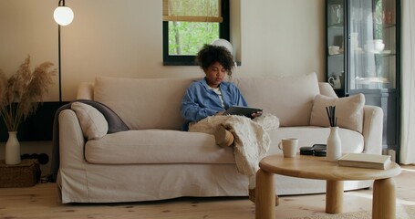 A woman in casual clothes uses a tablet sitting on the couch at home. She draws with a stylus