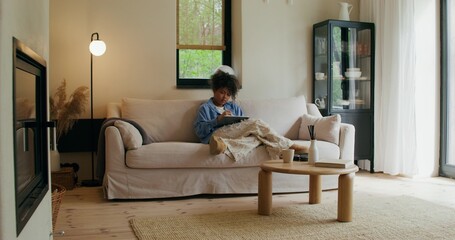 A woman in casual clothes uses a tablet sitting on the couch at home. She draws with a stylus