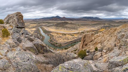 Obraz premium Panoramic view of a river bending through a rocky, desert landscape under a cloudy sky.