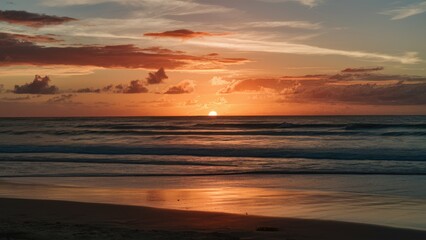Obraz premium Golden sunset reflecting on calm ocean waves near sandy beach with scattered clouds and vibrant orange, pink, and blue hues in the sky.
