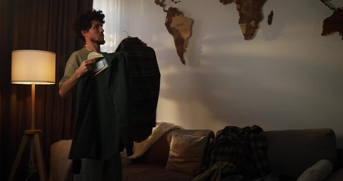 A brunette man with curly hair in a green T-shirt tries on different shirts and chooses one of them while getting dressed for work in the morning