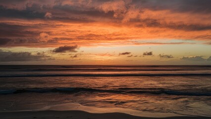 Obraz premium Vibrant orange and pink hues illuminate the ocean waves at sunset with dark clouds scattered across the sky creating a serene coastal scene.