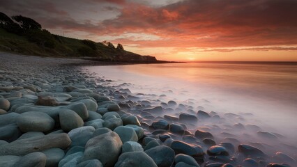Obraz premium Serene shoreline with smooth grey stones in the foreground, gentle waves reflecting orange and pink hues of sunset sky on the horizon.