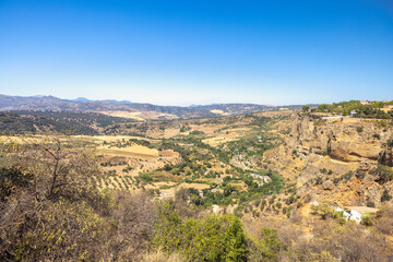 The mountainous land of Andalusia near Ronda town in Spain. Vast valley landscape viewed from a high vantage point, showcasing diverse terrain, vegetation and distant mountains under a clear blue sky.