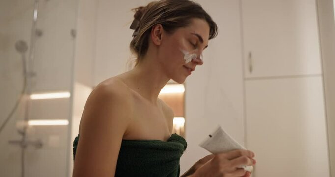 Happy brunette girl applies cream to her face after getting out of the shower in a modern bathroom