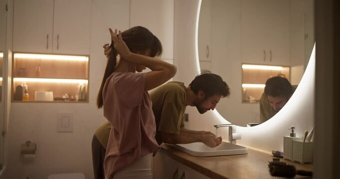 A guy hands over his face while his girlfriend ties her hair and does her hair in front of a mirror in a modern bathroom at home