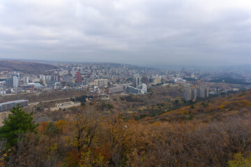 View from above on the western part of Tbilisi. Modern high rise residential buildings and offices. Autumn colors of grass, bushes and trees. Mountains in the background.