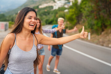 Cheerful interracial friends standing on road showing hitchhiking gestures. Tourism traveling