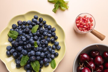 Blueberries, cherries and pomegranate seeds with mint leaves on beige background. Minimalistic and colorful composition of fresh fruits. Concept of healthy eating and summer