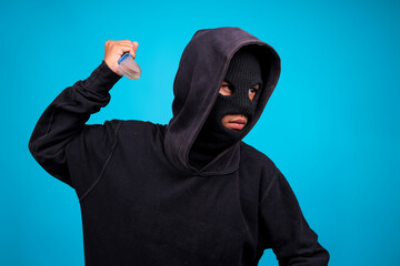 Man In A Black Hood Using Mask Stabbing With Knife Isolated On Blue Background 