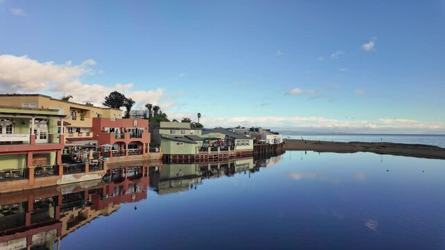 This is a high-quality panning clip of colorful buildings reflected in the water near the pier in Capitola, California, on a sunny day.