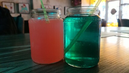 Two glass jars filled with colorful beverages, one pink and one blue, with bubbles and straws inside. The jars are placed on a wooden table in a dimly lit café or bar, with a blurred background featur