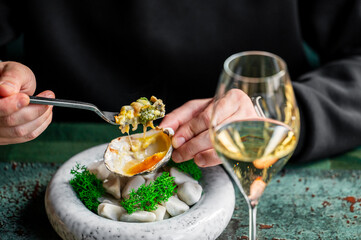 A close-up of a hand holding a fork with a delicious oyster, served on a decorative plate with green moss. A glass of white wine complements the dish in a stylish setting.
