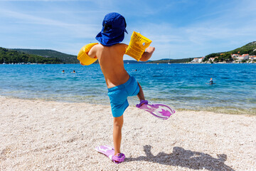 Back view cute little tanned boy kid in swimsuit sunscreen cap and fin funny walking dive in clear blue Mediterranean sea water on sunny summer day. Child have fun go snorkeling in flippers