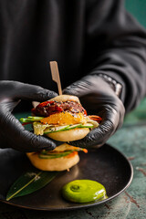 A close-up of a gourmet mini burger being held by a person in black gloves. The burger features layers of meat, vegetables, and sauces, served on a stylish dark plate with vibrant green sauce.