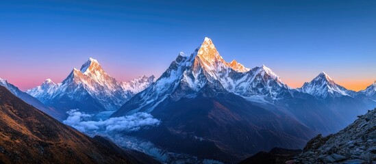 Obraz premium Majestic twilight panorama of snow-capped mountains bathed in golden and blue hues with a clear sky and soft clouds at the base of the range.