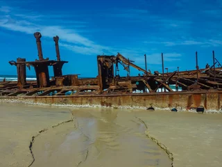 Fotobehang Rusty remains of SS Maheno shipwreck on Fraser Island's 75 Mile Beach against vibrant blue sky. Historic vessel and metal structures create dramatic silhouette on sandy shore with ocean waves © Chris