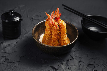 Crispy breaded shrimp served in a dark bowl with condiments and chopsticks on a textured black surface