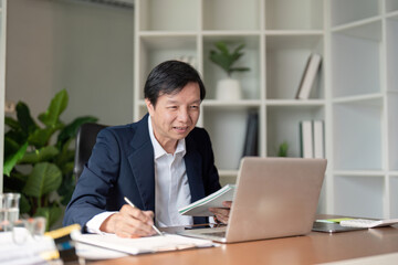 Elderly Asian businessman reviewing documents while taking notes on a laptop in a modern office.