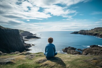 Young boy enjoying peaceful moment, taking in serene view of blue ocean from grassy clifftop under cloudy sky