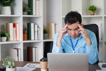 Stressed Asian businessman focusing on a laptop, dealing with challenges at his desk in a modern...