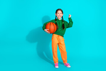 Adorable young girl holding a basketball in casual attire against a vibrant turquoise background showcasing playful and trendy fashion