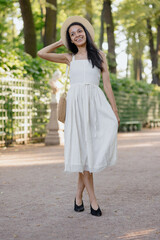 carefree African American woman in white dress and straw hat walks in park and poses against background of green hedge. daytime for relaxation. beautiful female fashion model, sunlight summer time
