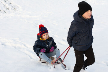 A child pulls a sled carrying a younger sibling across a snow-covered field. Both children are...
