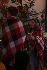 Obraz premium Two children wrapped in red and white plaid blankets, near a Christmas tree adorned with lights and ornaments. One holds a warm beverage. A festive, winter scene.