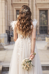 Modern young bride in wedding dress from behind with glam wavy hairstyle with crystal haircomb outdoors. No face woman with blond curly long hair.