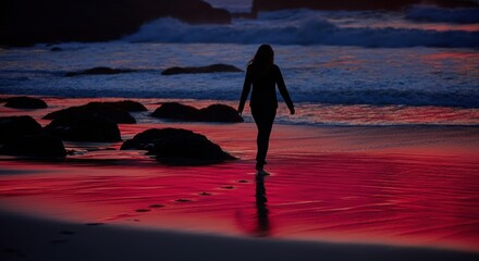 Silhouette of a woman walking along a beach at sunset, creating footprints in the sand
