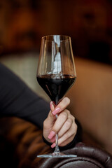 A close-up of a woman's hand holding a glass of red wine. The elegant glass is filled with dark wine, set against a blurred background of a cozy interior. Ideal for lifestyle and dining themes.