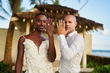 Bride in white dress, groom in bold outfit show rings at lgbtq wedding in tropical villa. Happy...