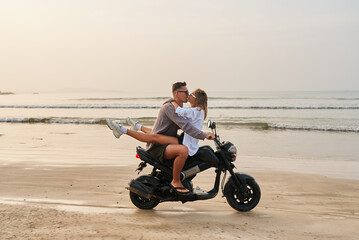 Young couple rides motorbike on beach. Lovers on motorcycle kiss at ocean shore. Romantic honeymoon travel. Man drives scooter with girlfriend. Adventure, passion, vacation.