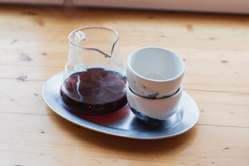 Glass server with black filter coffee inside and ceramic cup stand on wooden cafeteria table, day light from window. Making filtered coffee in coffee shop. Pour over method of alternative brewing, v60