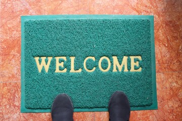 A green welcome doormat with feet wearing black shoes. Interior and object concept.