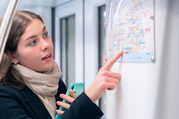  Focused young woman in a black coat and beige scarf checking a subway map while holding her...