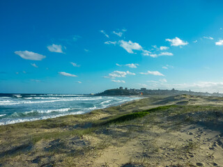 Dramatic coastal landscape of Newcastle Beach, Australia, featuring turbulent ocean waves crashing against sandy shores. Grassy sand dunes frame the city skyline silhouette. scattered white clouds