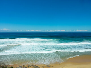 Turquoise waves crash onto golden sandy beach under vibrant blue sky, Newcastle Beach, Australia. Crystal-clear ocean waters create dynamic white foam patterns. Pristine shoreline on sunny summer day