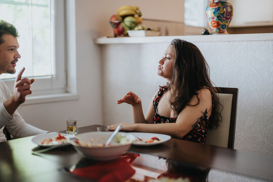 Young couple engaging in an animated conversation while sitting across each other at a dining table. The scene conveys a mix of emotions as they discuss over a meal.