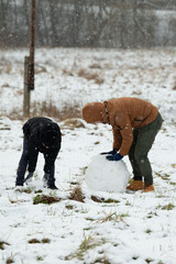 On a snowy day, two individuals work together to construct a snowman. They are bundled in winter...